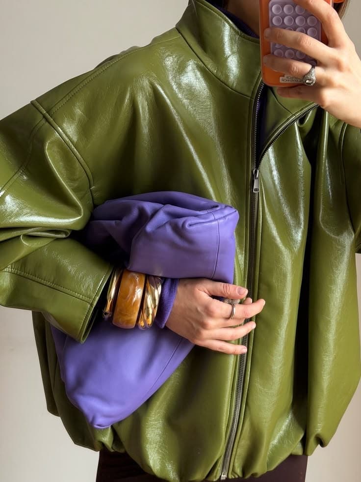 A person in a glossy olive-green leather jacket holding a lilac bag, photographing themselves in a mirror